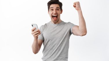 Happy man holds smartphone and celebrating, winning online prize, shouts yes and rejoicing, triumphing from success, looks satisfied, white background.