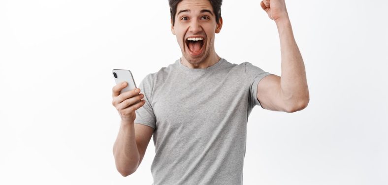 Happy man holds smartphone and celebrating, winning online prize, shouts yes and rejoicing, triumphing from success, looks satisfied, white background.